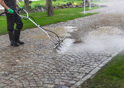 person using a power washer on a cobblestone driveway surrounded by grass and trees cleaning the surface with high pressure water